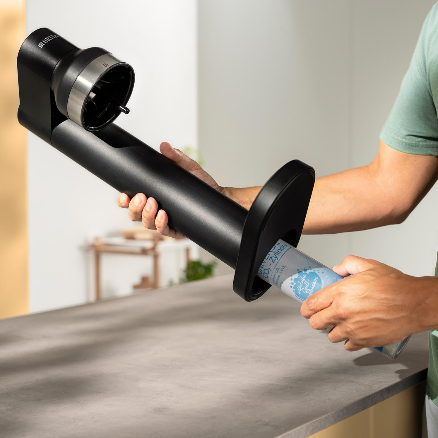 Osoba wkłada cylinder CO2 do czarnego urządzenia. Urządzenie ma markę BRITA. Scena ma miejsce w pomieszczeniu na szarym blacie.
