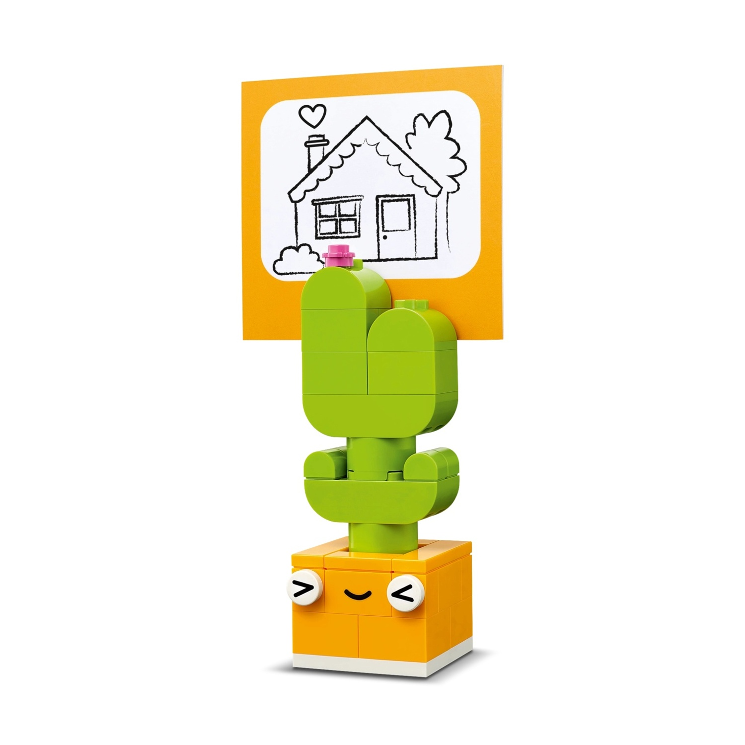 Un giocattolo cactus Lego con un disegno di una casa. Il cactus è verde, in un vaso arancione. Il disegno della casa è su sfondo arancione.