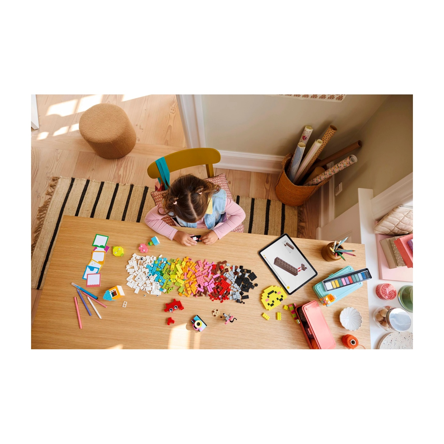 Una ragazza è seduta a un tavolo con blocchi colorati, un tablet e materiale artistico. Uno sgabello e una sedia sono vicini, con un tappeto sotto.