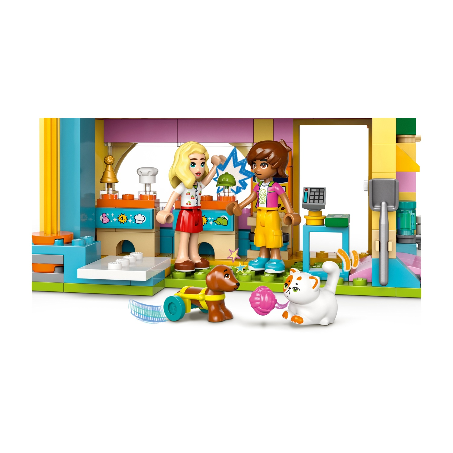 Set Lego. Due ragazze Lego, un cane e un gatto in una cucina. Una ragazza saluta. Il gatto gioca con una palla rosa.