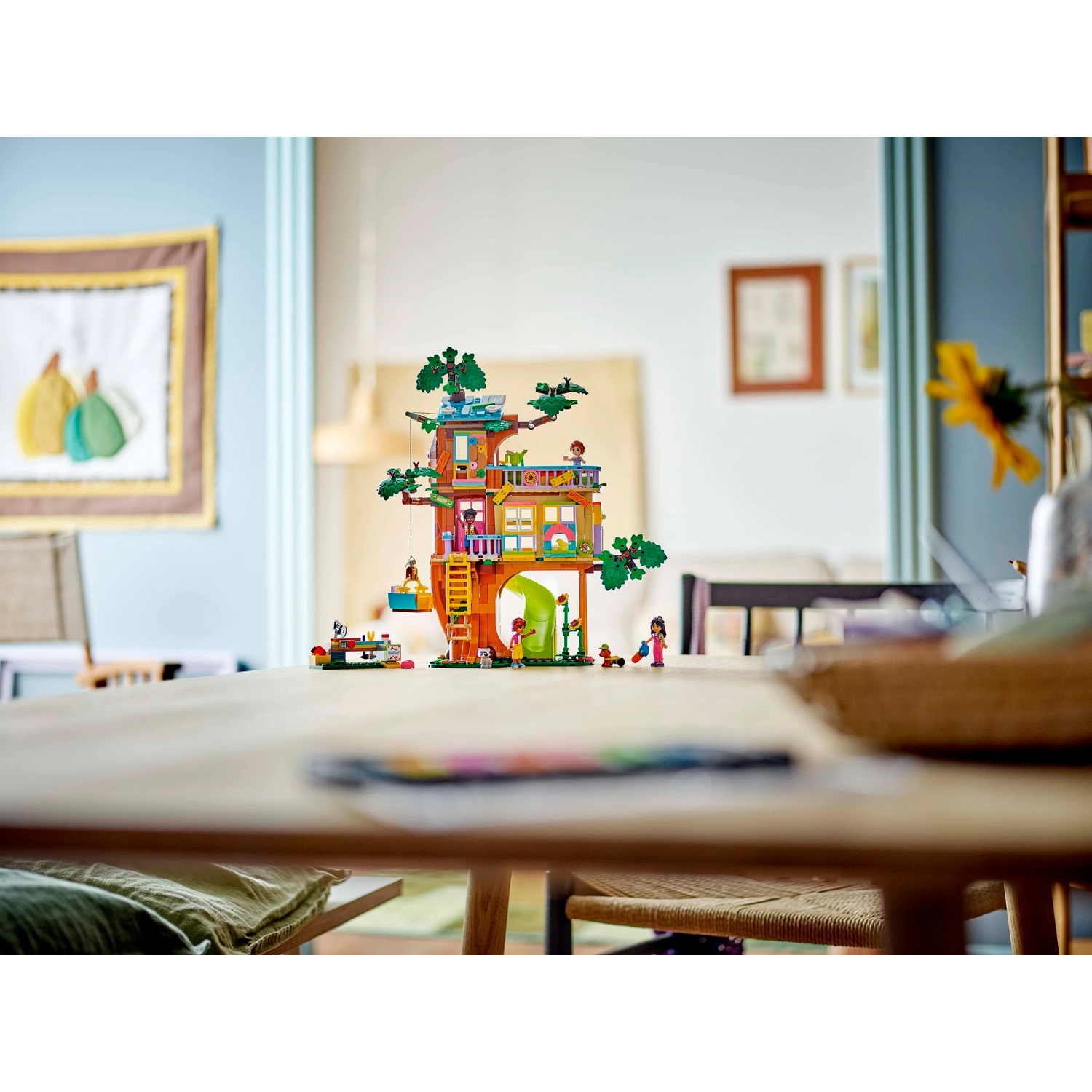Un set di gioco Lego di una casa sull'albero è appoggiato su un tavolo di legno. La casa sull'albero ha più livelli e personaggi. Sfondo con dipinto e fiore.