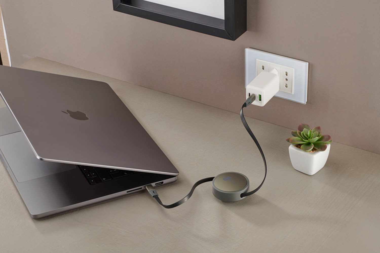 Una MacBook está conectada a un cargador. Hay una planta pequeña en una maceta blanca sobre la mesa, cerca del enchufe.