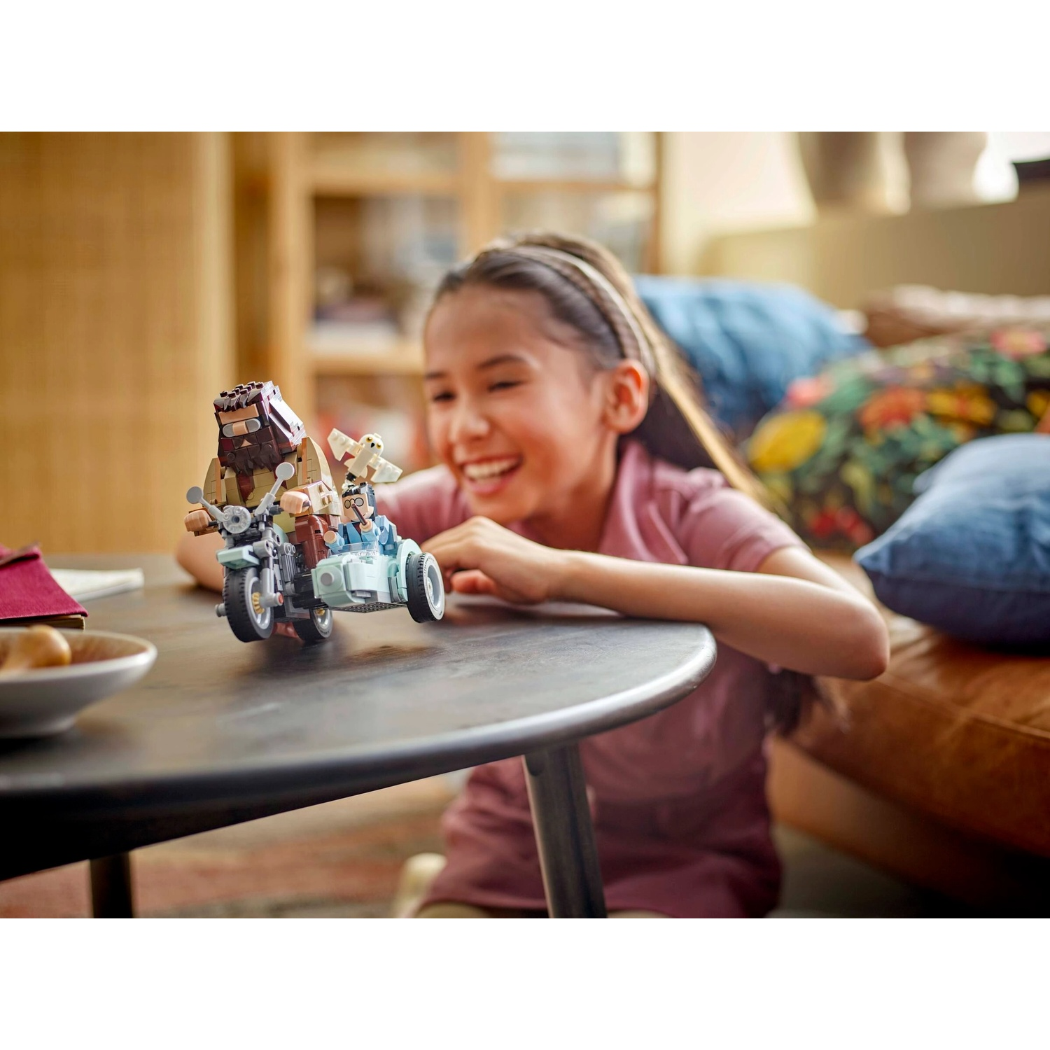 Una ragazza sorride a una moto Lego con sidecar. La moto ha un personaggio Lego e un piccolo gufo bianco. Un tavolo è a fuoco.