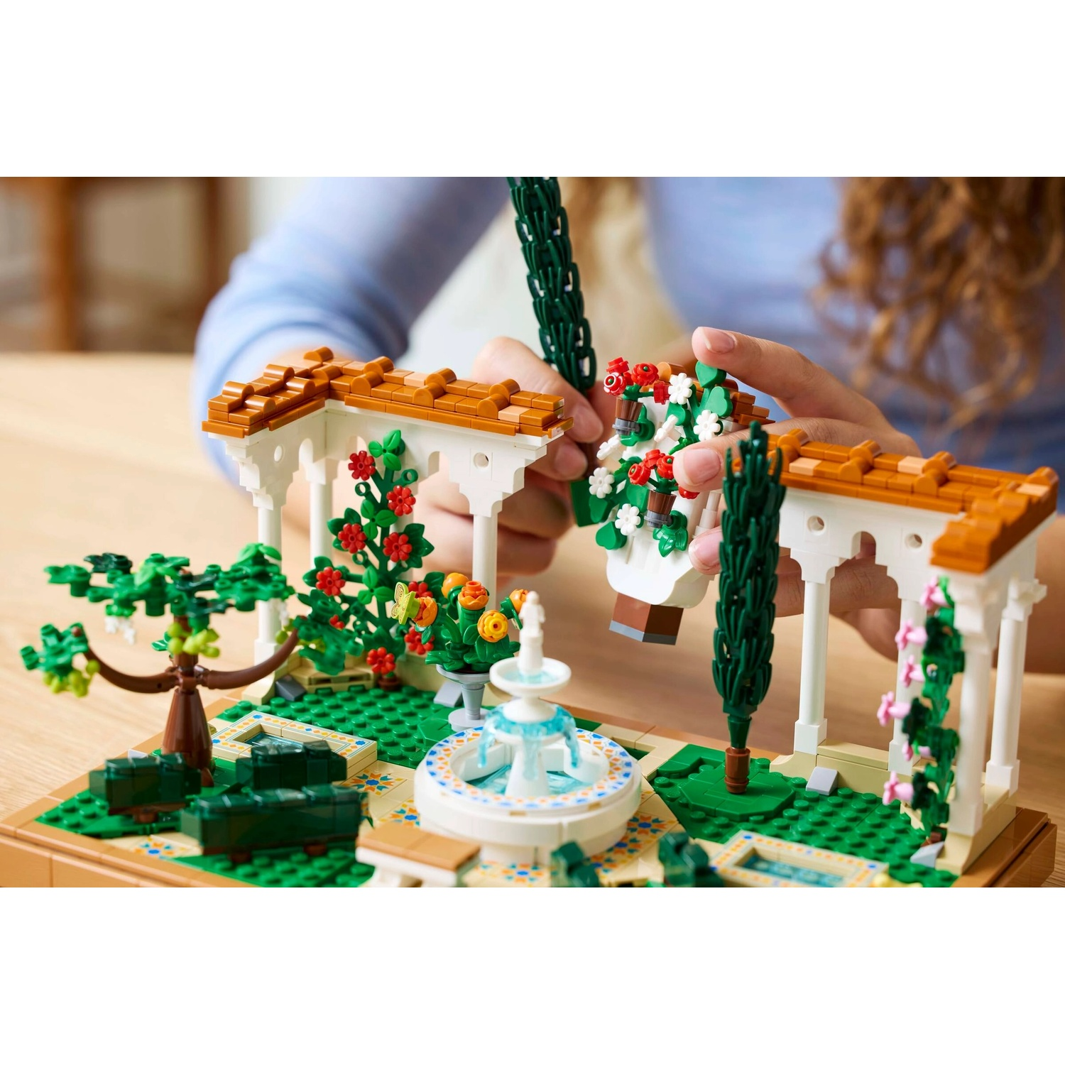 Una persona sta assemblando una scena del giardino Lego. Presenta una fontana, archi, alberi e composizioni floreali, con una base verde.