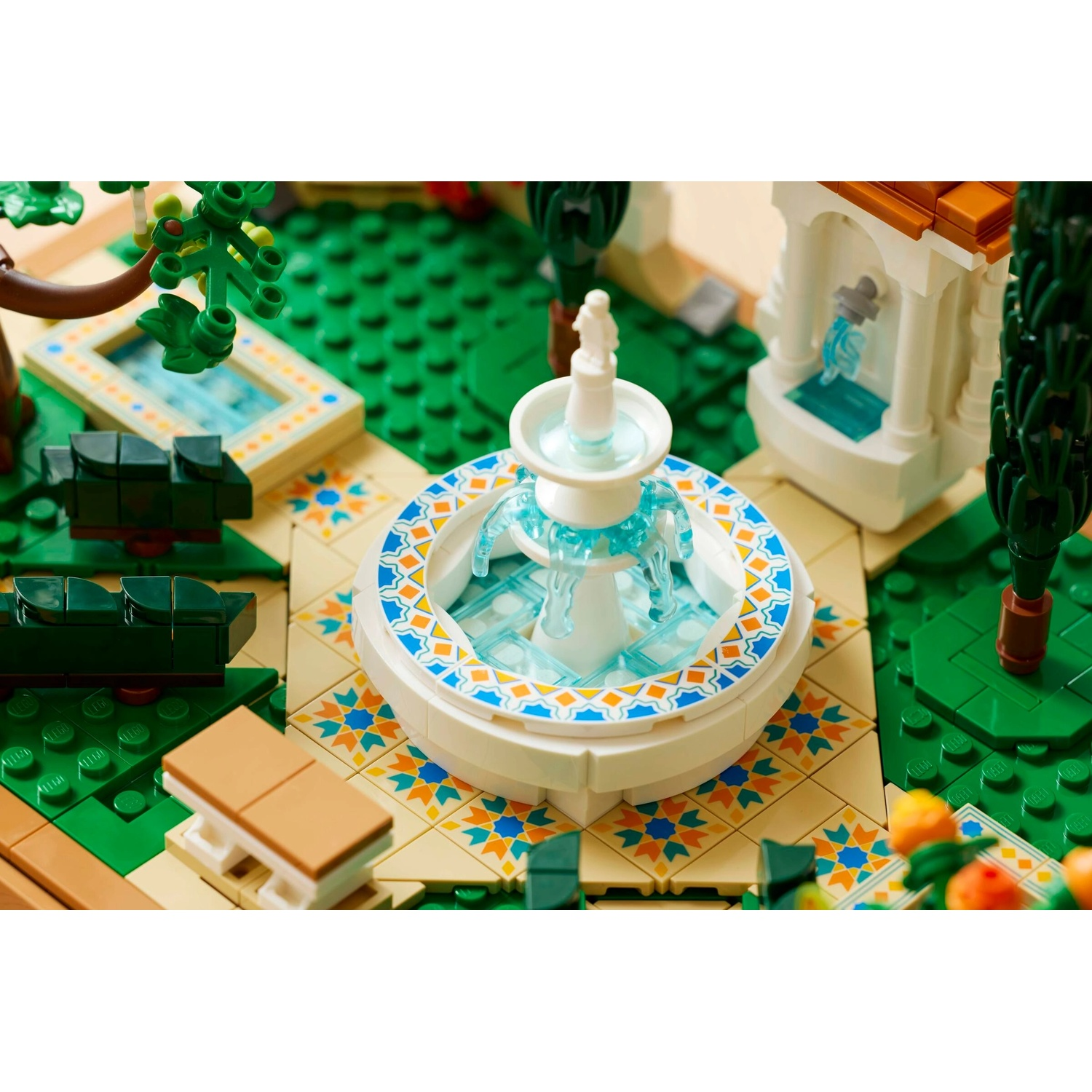 Una fontana Lego con acqua blu è circondata da un giardino. C'è una piccola piscina, alberi e piastrelle decorative. Edificio bianco sullo sfondo.