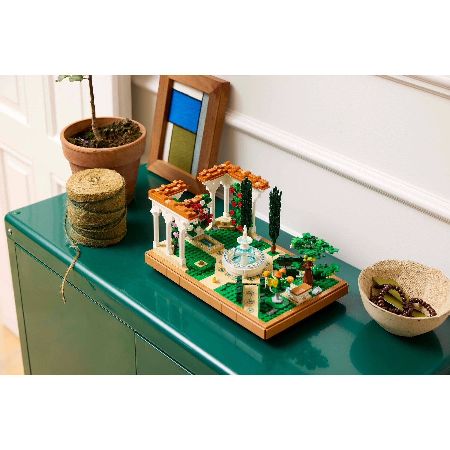 Una scena Lego di un giardino con fontana, gazebo e vegetazione su un mobile verde. Una pianta, uno spago e una ciotola sono vicini.