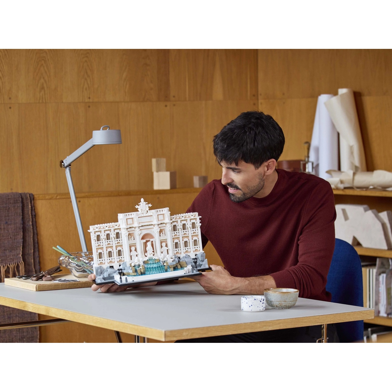 Un uomo con un maglione bordeaux esamina un modello di edificio. Il modello è su un tavolo con una lampada e delle ciotole.