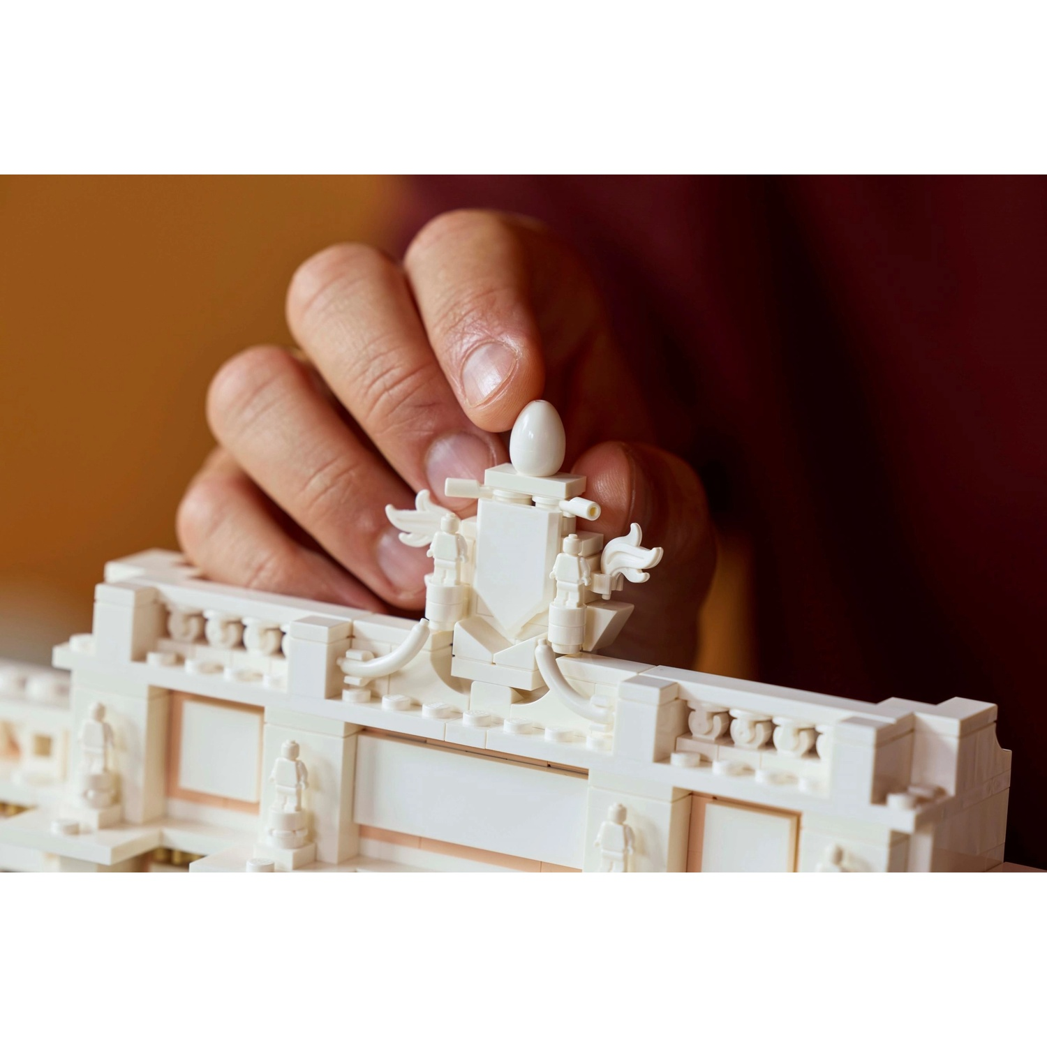 Una mano posiziona un pezzo Lego bianco su un modello Lego di un edificio con statue. Lo sfondo è di colore rosso sfocato.