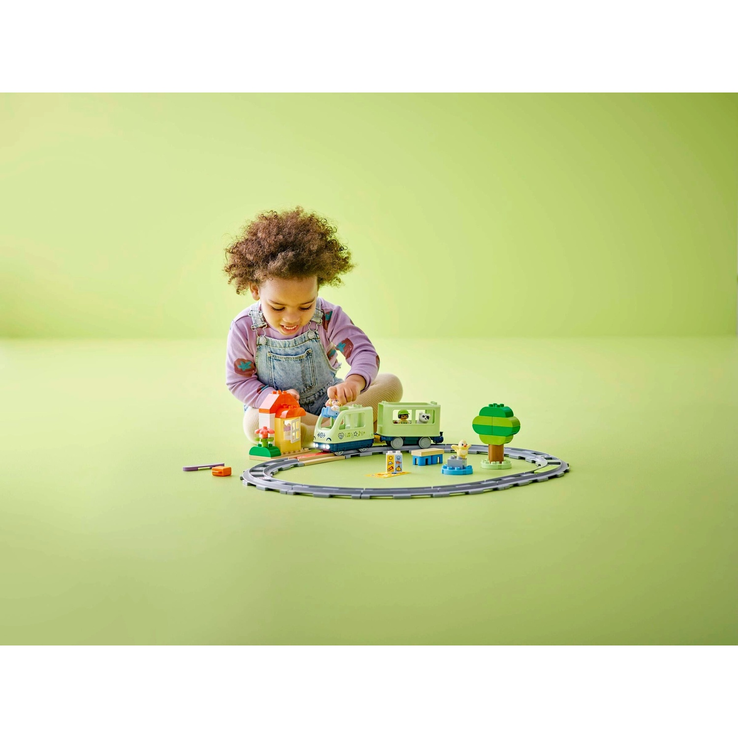 Un bambino gioca con un trenino giocattolo. La scena comprende un treno, un binario, blocchi di costruzione e un albero, il tutto su sfondo verde.
