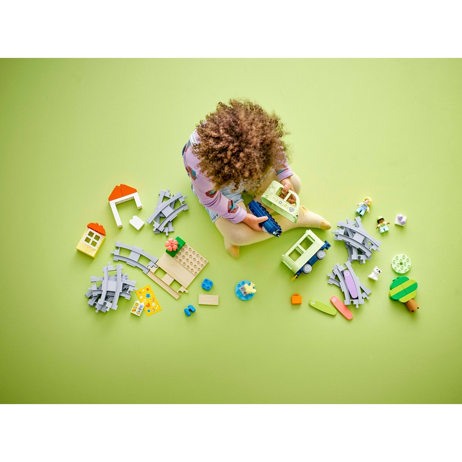 Un bambino è seduto circondato da pezzi di un set di trenini giocattolo su una superficie verde. Ci sono vagoni, binari, figure, una casa e alberi.