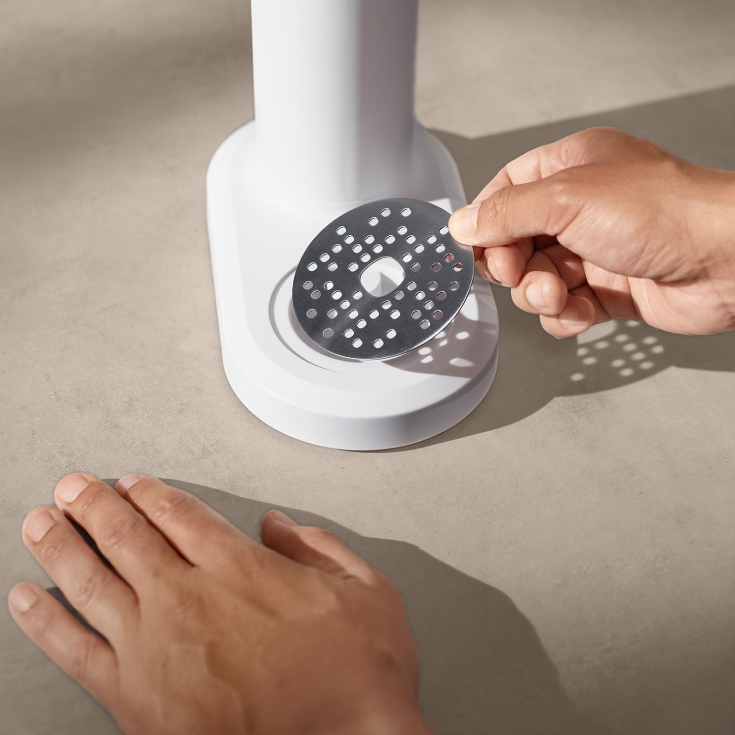 Dłoń trzyma metalową tarczę z otworami, umieszczając ją na białym urządzeniu kuchennym na beżowej powierzchni.