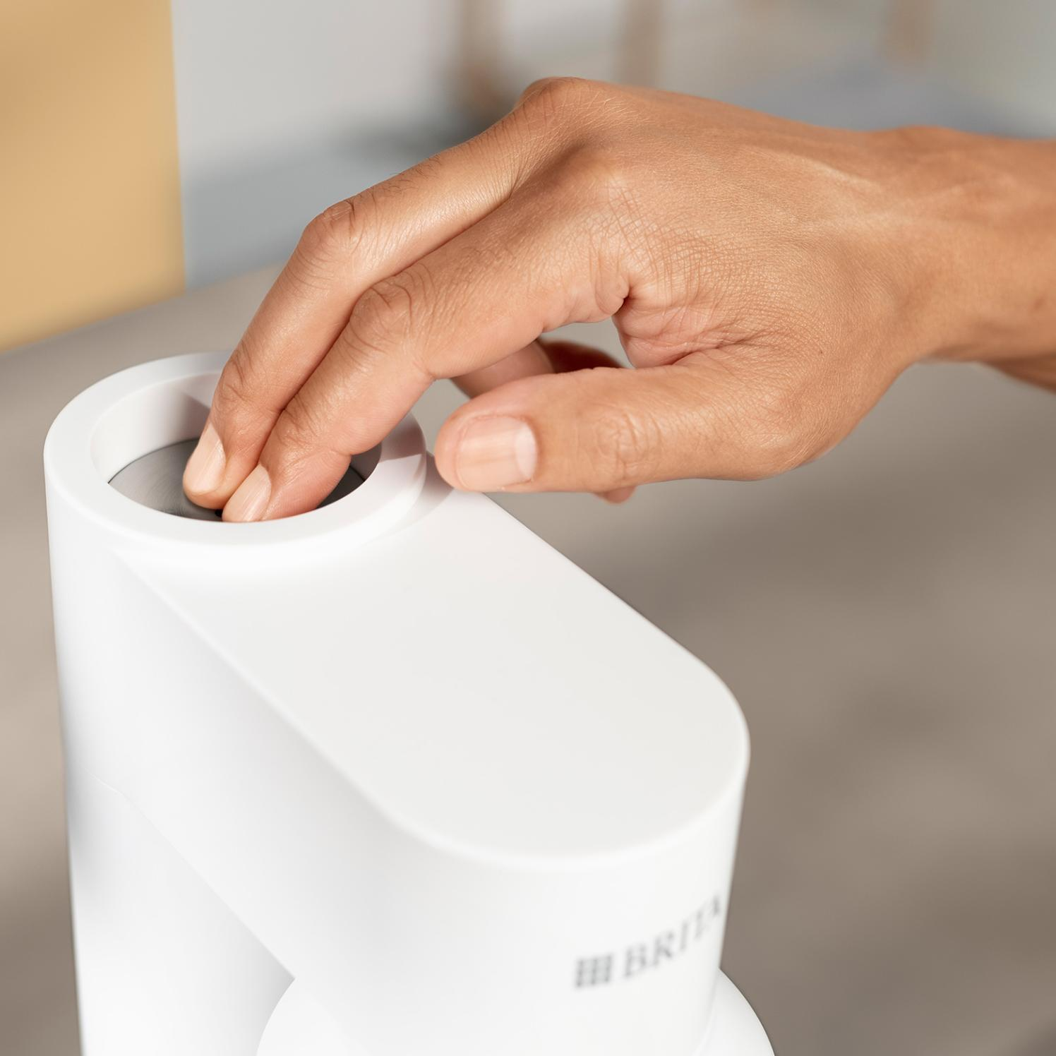 Ręka dotyka białe urządzenie. Urządzenie ma okrągły otwór. Tło to jasnoszara powierzchnia.