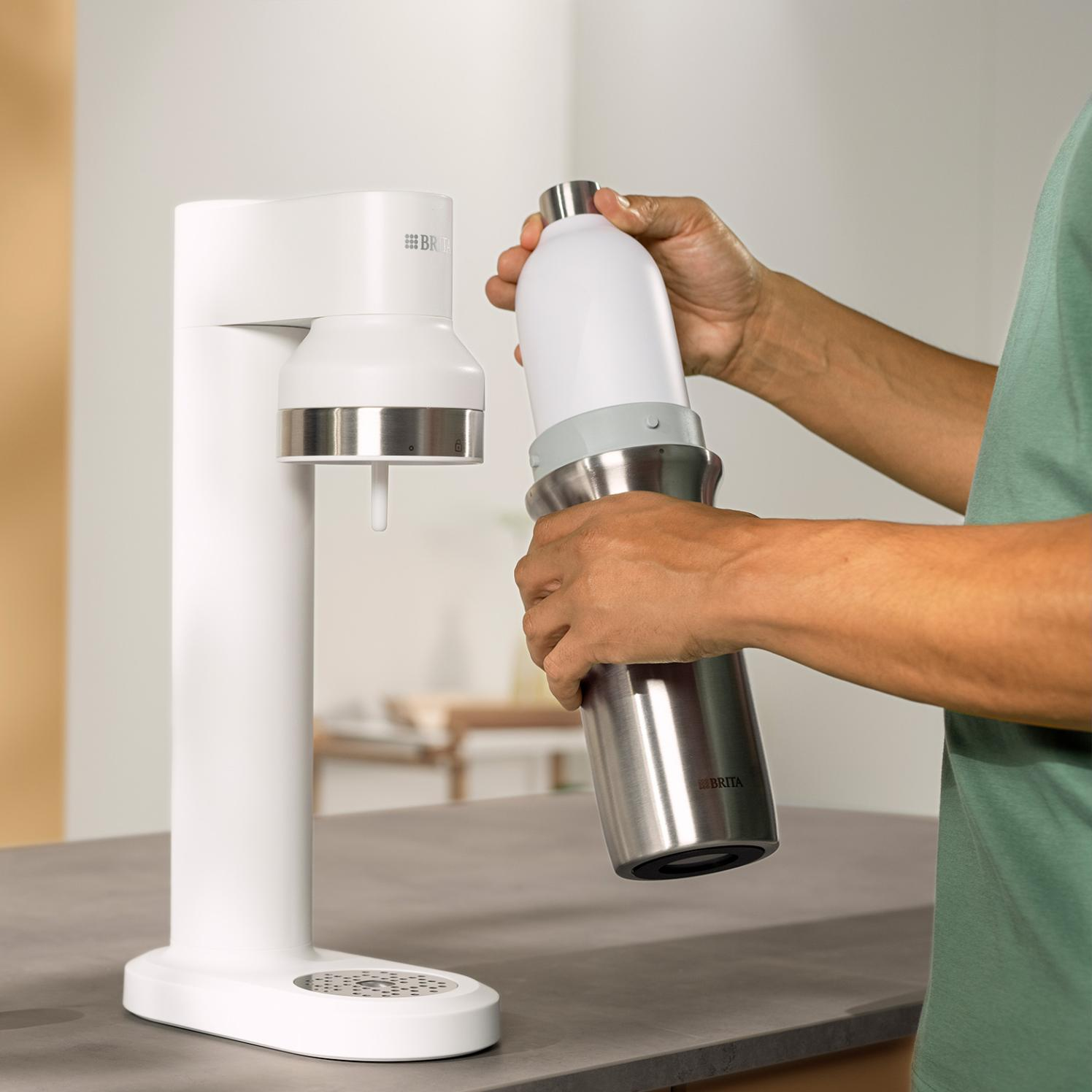 Osoba trzymająca butelkę z wodą i białe urządzenie do wody gazowanej na szarym blacie. Osoba ma na sobie zieloną koszulę.