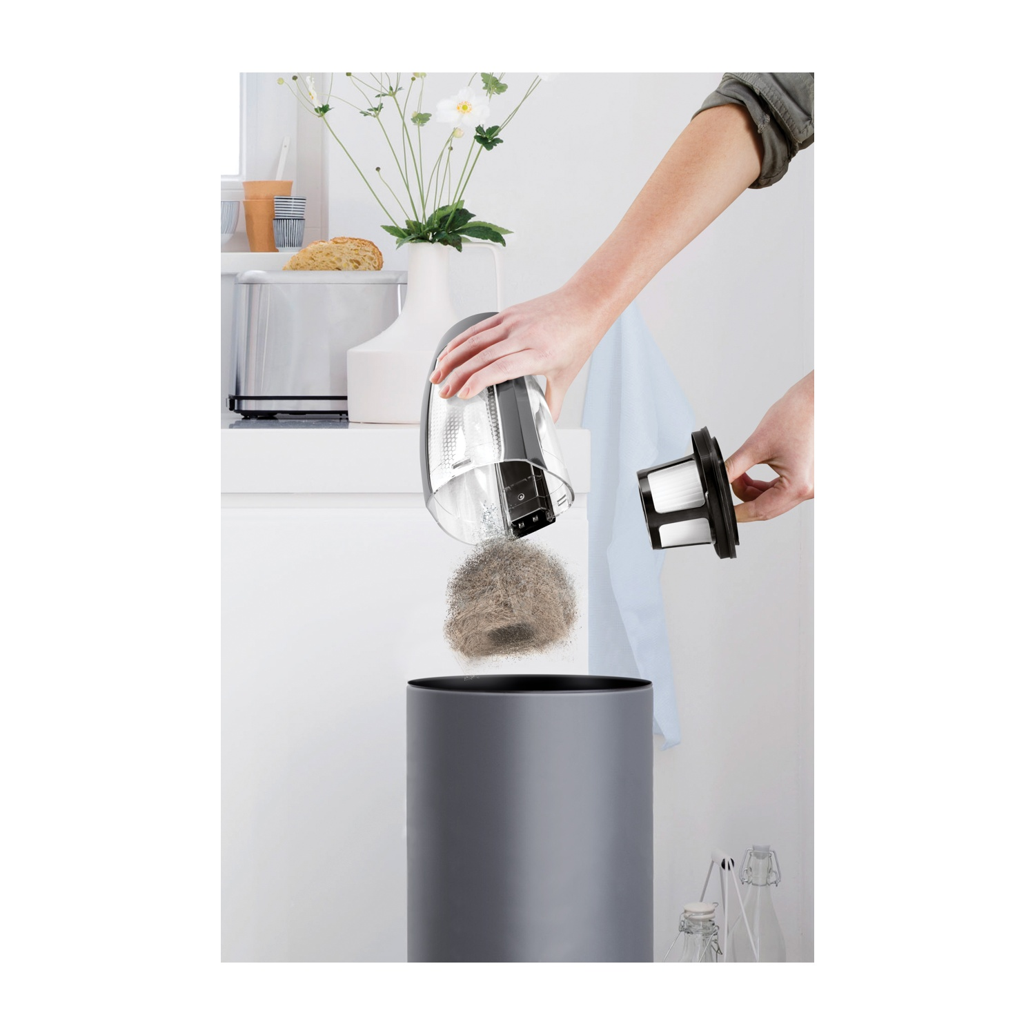 Una persona sta svuotando il contenitore della polvere di un aspirapolvere in un cestino grigio. Un filtro e polvere vengono rimossi.