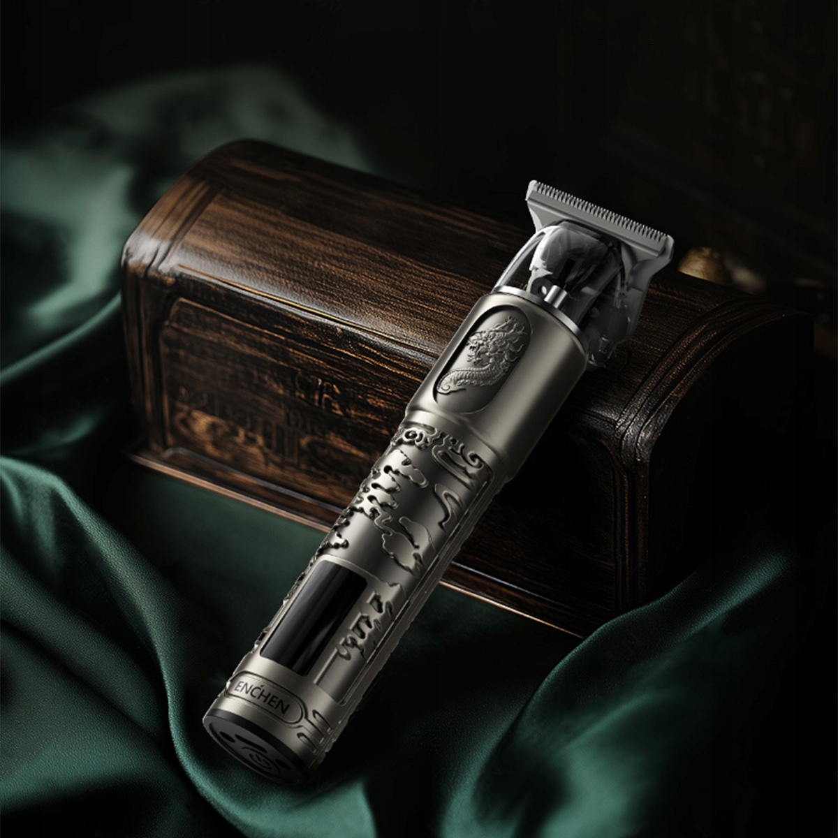 Srebrna maszynka do włosów ze skomplikowanym wzorem, umieszczona na zielonej tkaninie obok drewnianej skrzynki. Trymer ma motyw smoka.