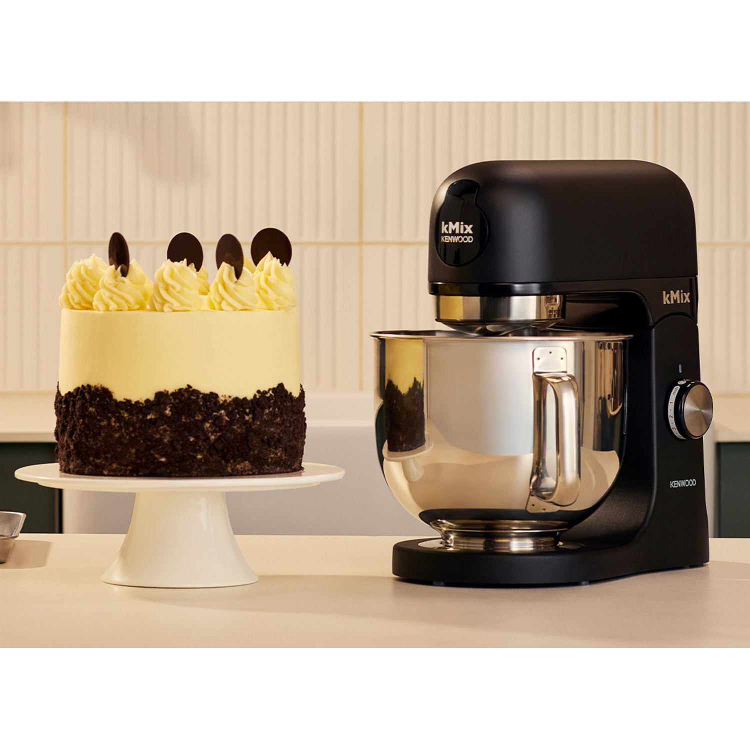 A black Kenwood Kmix mixer sits next to a cake on a white stand. The cake has frosting and chocolate decorations.