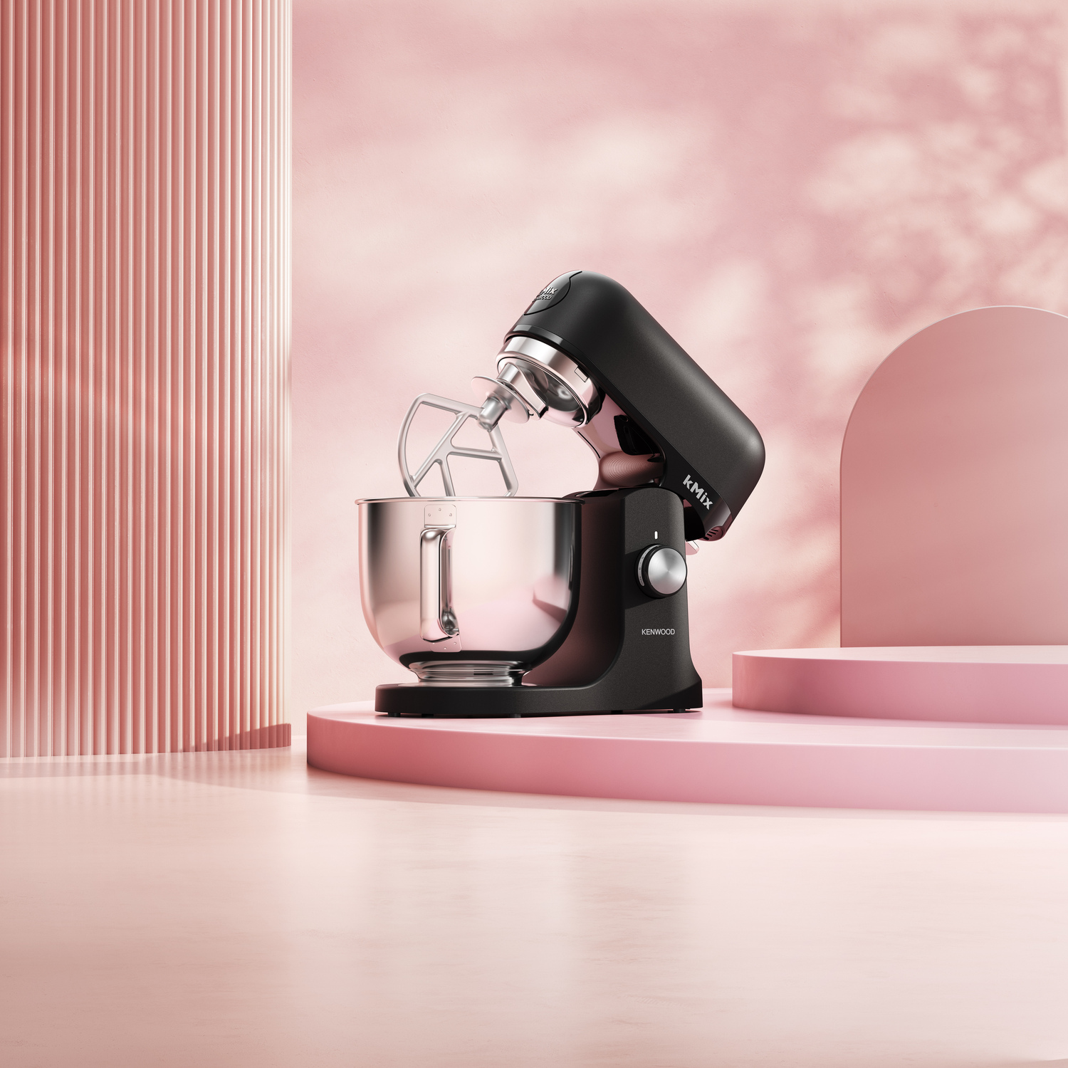 A black Kenwood Kmix stand mixer on a pink platform. A silver mixing bowl and beater are attached. Pink background with architectural elements.