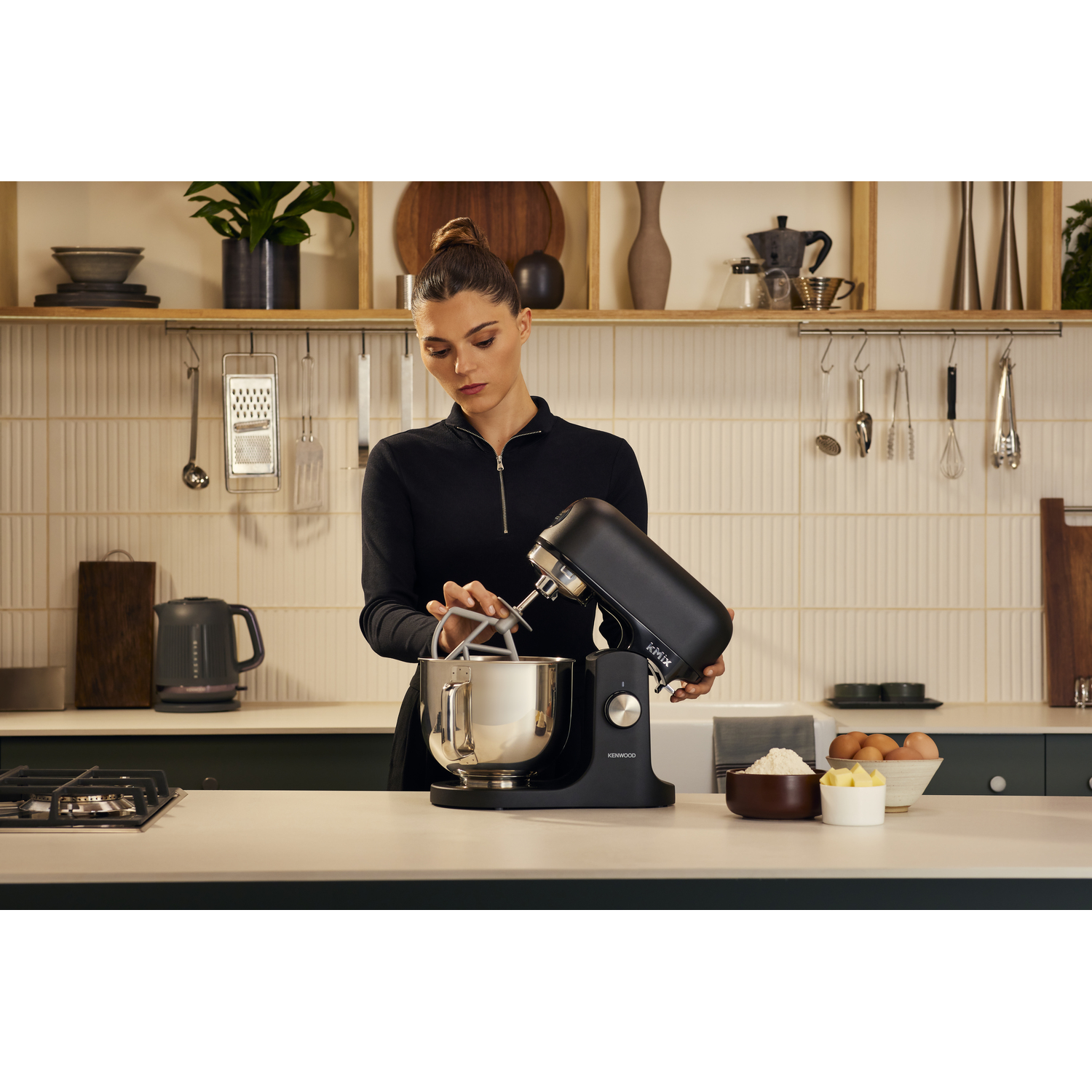 A woman using a black stand mixer in a kitchen. Ingredients like flour, butter, and eggs are on the counter.