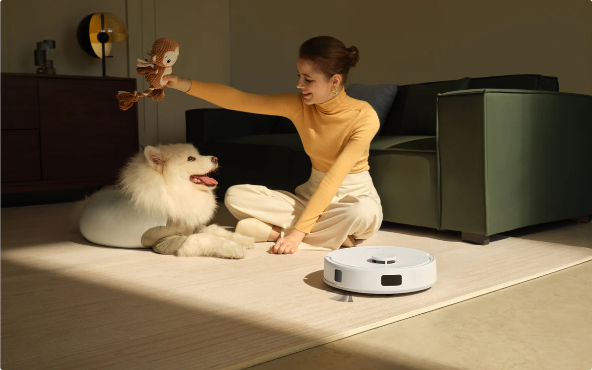 Femme avec chien et jouet, près d'un aspirateur robot. Femme en haut jaune, chien blanc. La pièce a un canapé et une lampe.