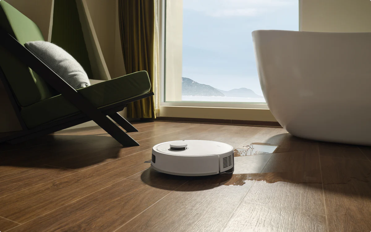 Un aspirateur robot blanc nettoie un plancher en bois. Une chaise verte, une baignoire blanche et une fenêtre avec vue sur la montagne sont visibles.