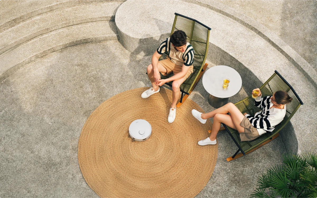 Deux personnes se détendent en plein air. L'un boit, l'autre est assis. Un aspirateur robot se trouve sur un tapis entre eux.