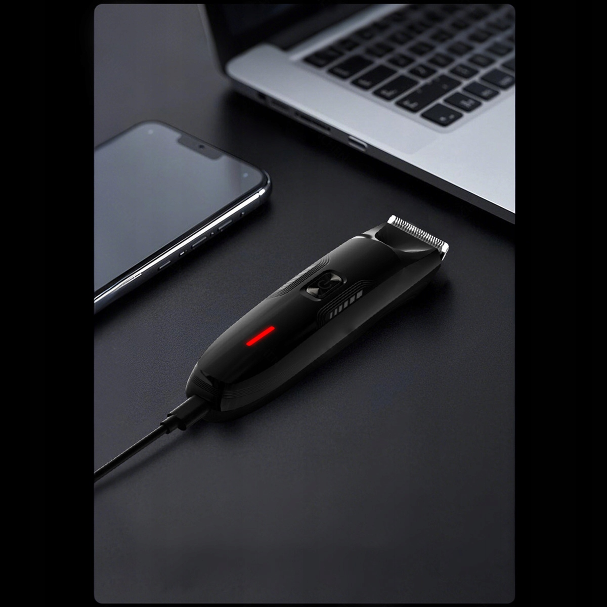 Czarna golarka elektryczna z czerwoną lampką kontrolną, telefon i laptop na ciemnej powierzchni.