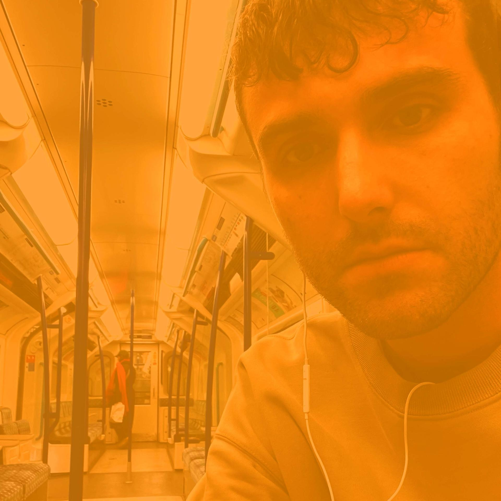 Egy férfi sárga pulóverben a metrón, a kamerába néz. Háttérben egy másik utas, piros sállal.