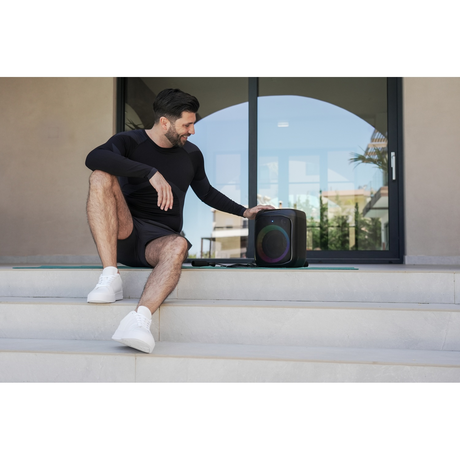 Un uomo in abbigliamento sportivo nero è seduto sui gradini accanto a un altoparlante. Sorride, guardando l'altoparlante. Lo sfondo mostra una porta a vetri.