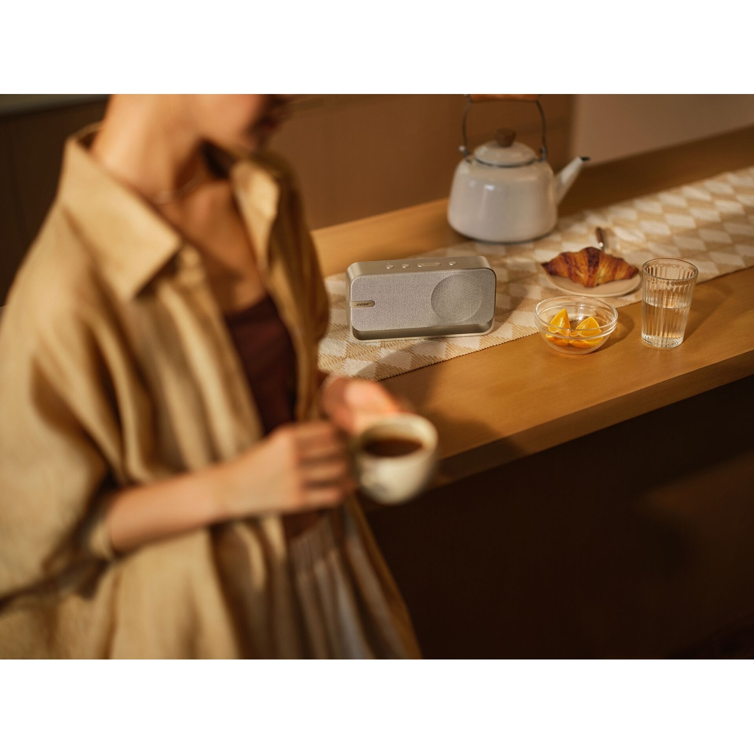 Una donna tiene in mano il caffè vicino a un tavolo con un altoparlante, un croissant, frutta, un bicchiere d'acqua e un bollitore.