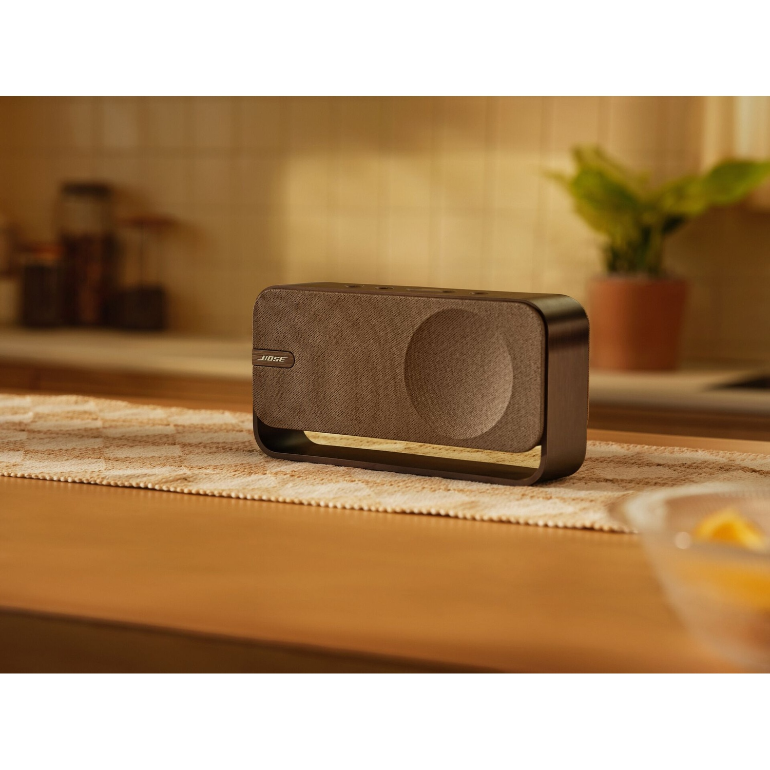 Un altoparlante Bose su un tavolo di legno. È marrone, con un design circolare. Dietro, una pianta in vaso e un'ambientazione da cucina.