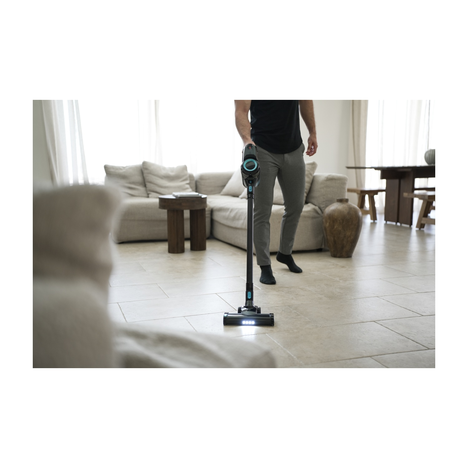 Una persona pulisce con l'aspirapolvere un pavimento piastrellato con un aspirapolvere senza fili nero e verde acqua. Sullo sfondo un divano beige, un tavolino e un tavolo di legno.