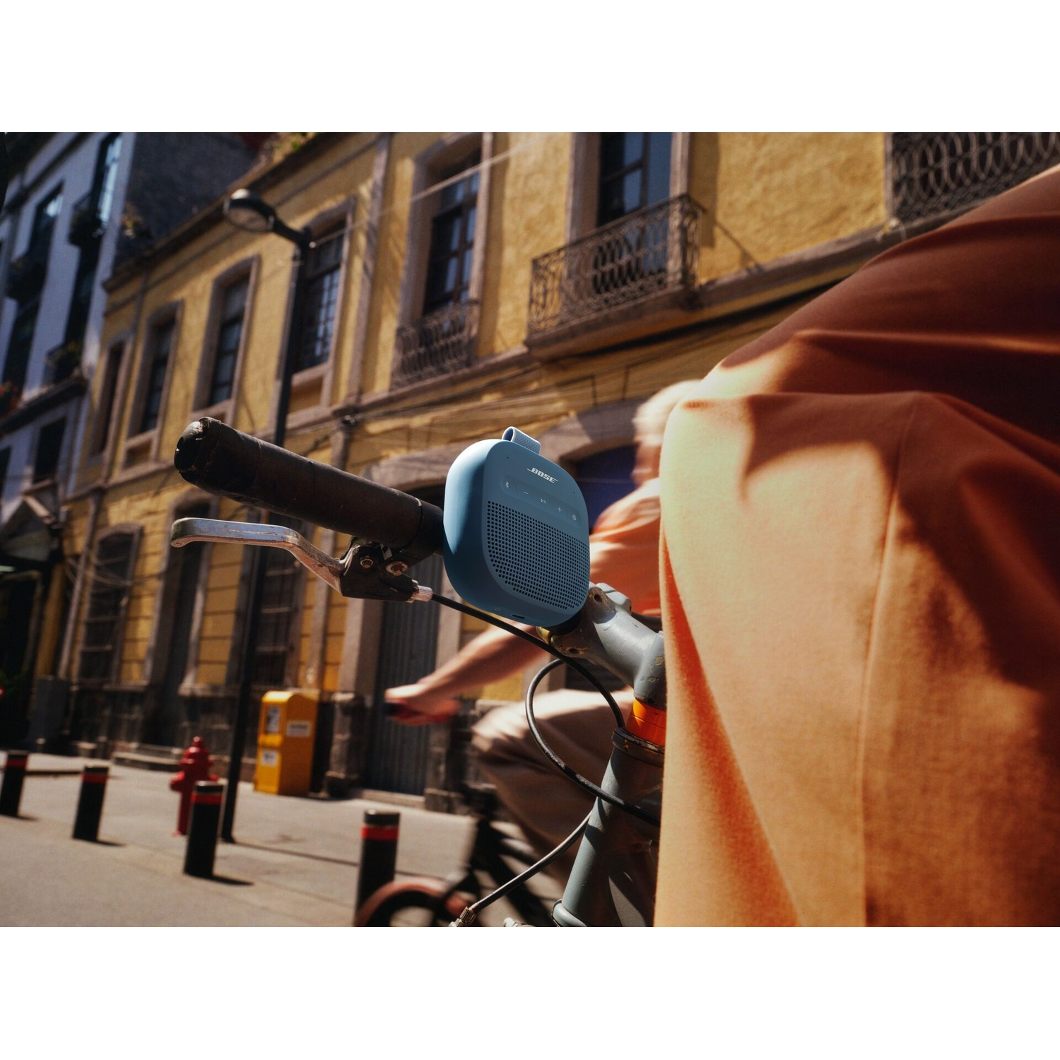 Una persona in bicicletta con un altoparlante blu attaccato. Edificio giallo sullo sfondo. Focus sull'altoparlante e sugli abiti della persona.