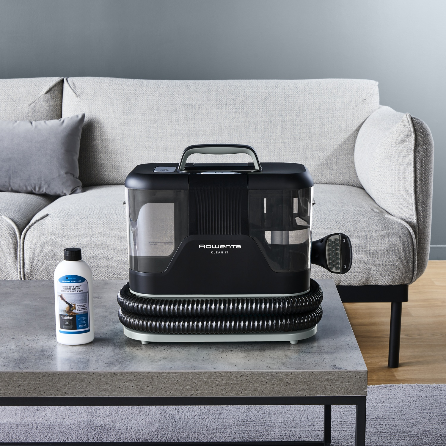 Un nettoyeur Rowenta Clean It noir et transparent sur une table, à côté d'une bouteille de solution de nettoyage. Un canapé gris en arrière-plan.