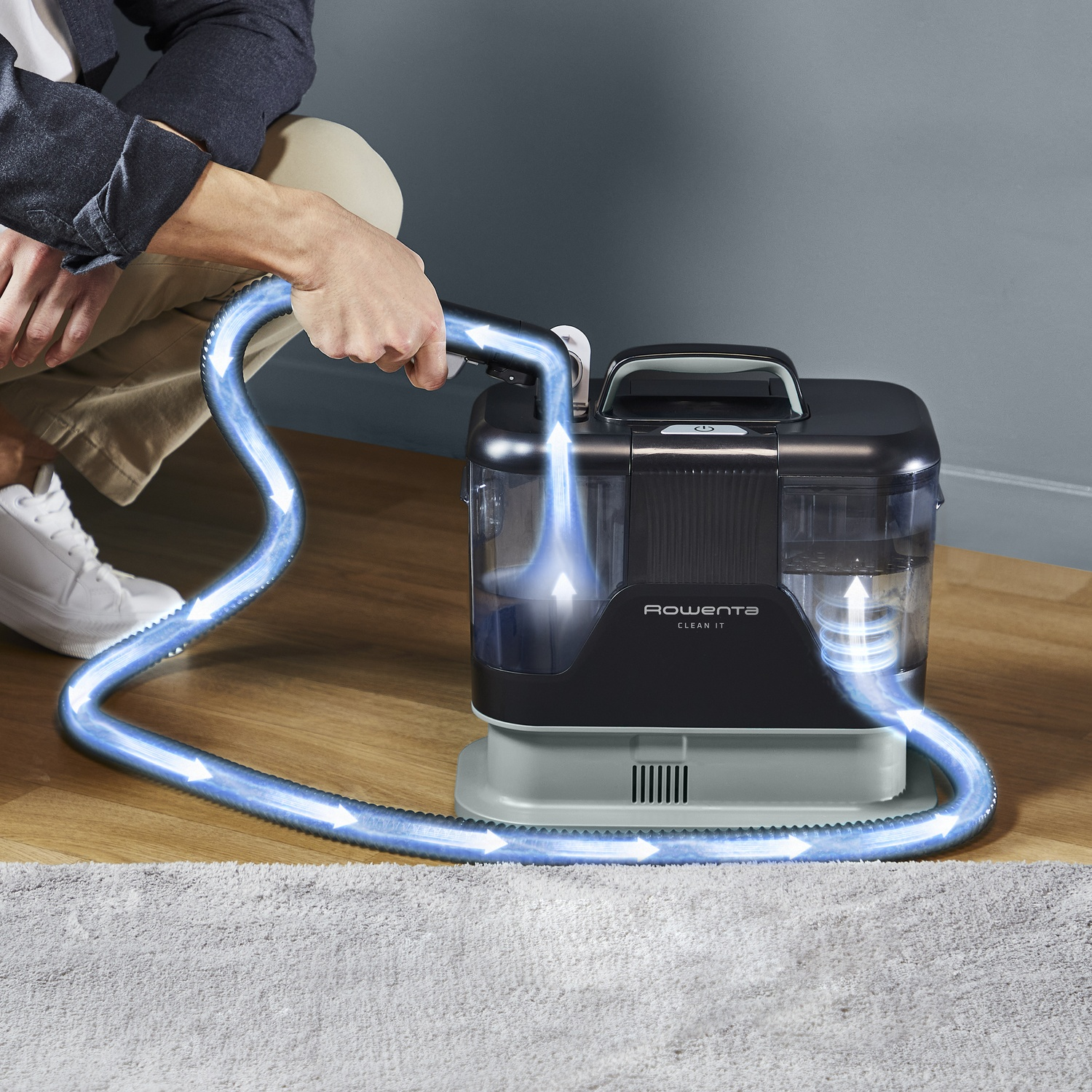 Une personne utilise une machine Rowenta Clean It pour nettoyer un tapis. Des flèches indiquent le flux d'eau de la machine via un tuyau.