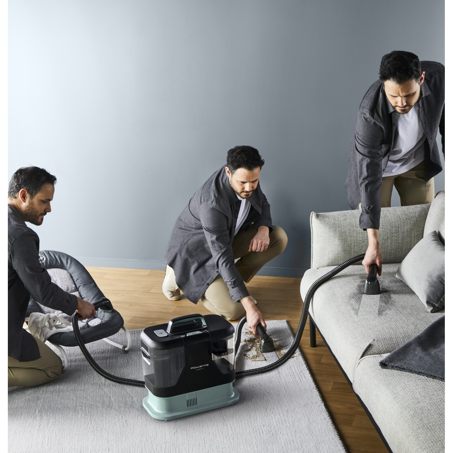 Trois hommes utilisent un nettoyeur de tapis sur un canapé, un tapis et un transat. Machine au milieu. Murs gris et tapis gris clair.