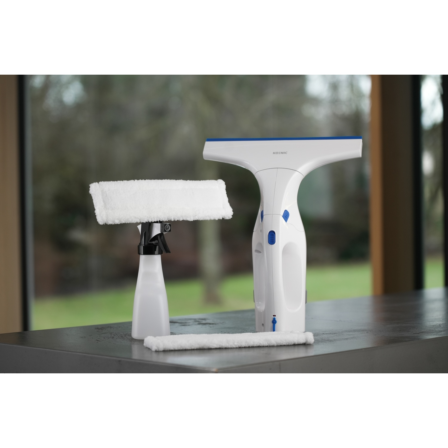 Un kit per la pulizia dei vetri su una superficie grigia. Include un flacone spray, un aspiragocce e un tampone per la pulizia. Lo sfondo mostra alberi.