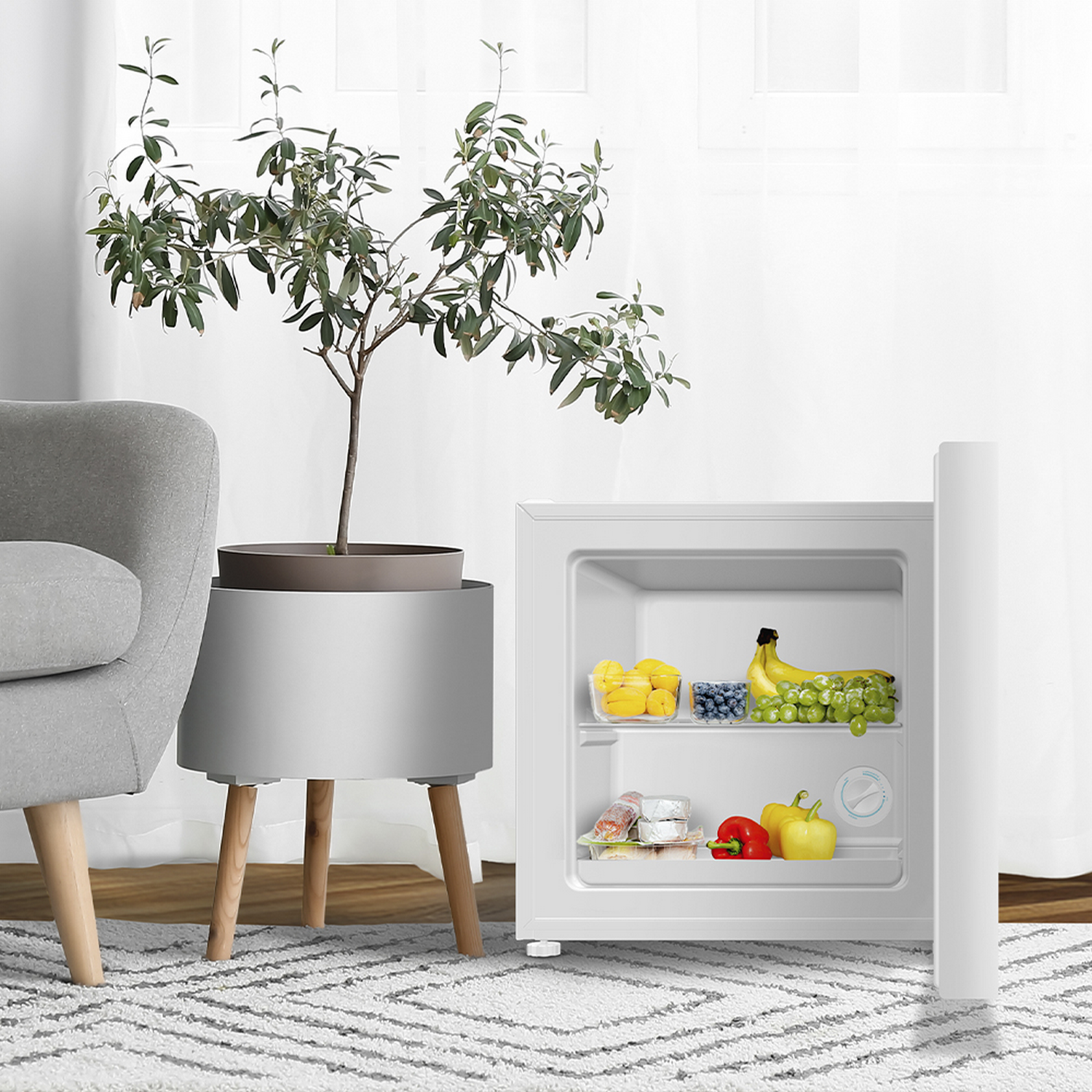 Un mini-refrigerador lleno de frutas y verduras. Una planta en maceta y una silla gris están al lado. Hay una alfombra en el suelo.