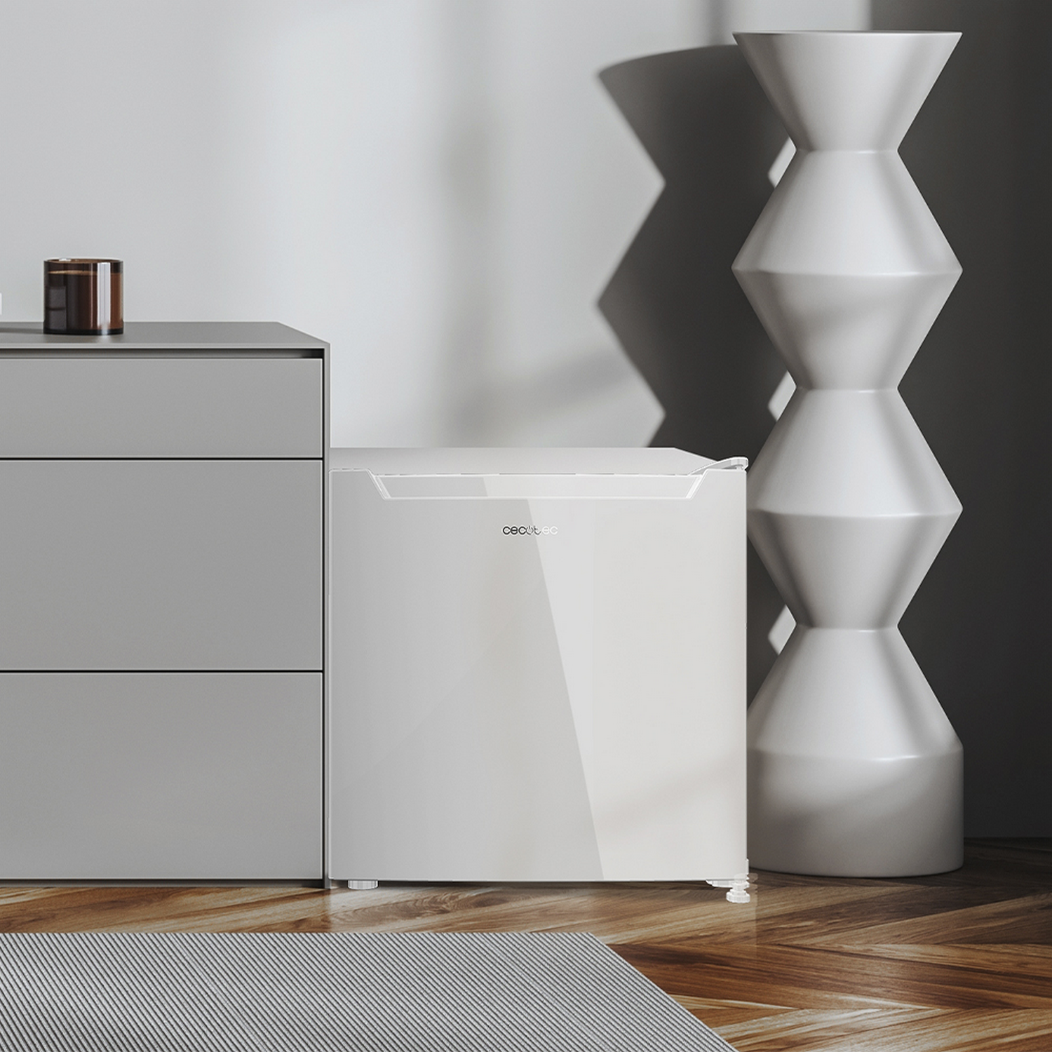 Un pequeño refrigerador blanco junto a un gabinete gris y un jarrón blanco decorativo. Piso de madera con una alfombra en primer plano.
