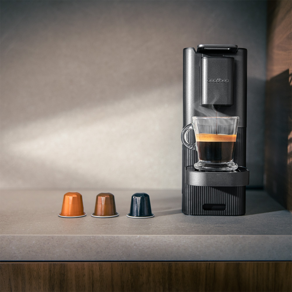 Una cafetera negra con un vaso de espresso. Hay tres cápsulas de café al frente. El vapor sube del espresso.