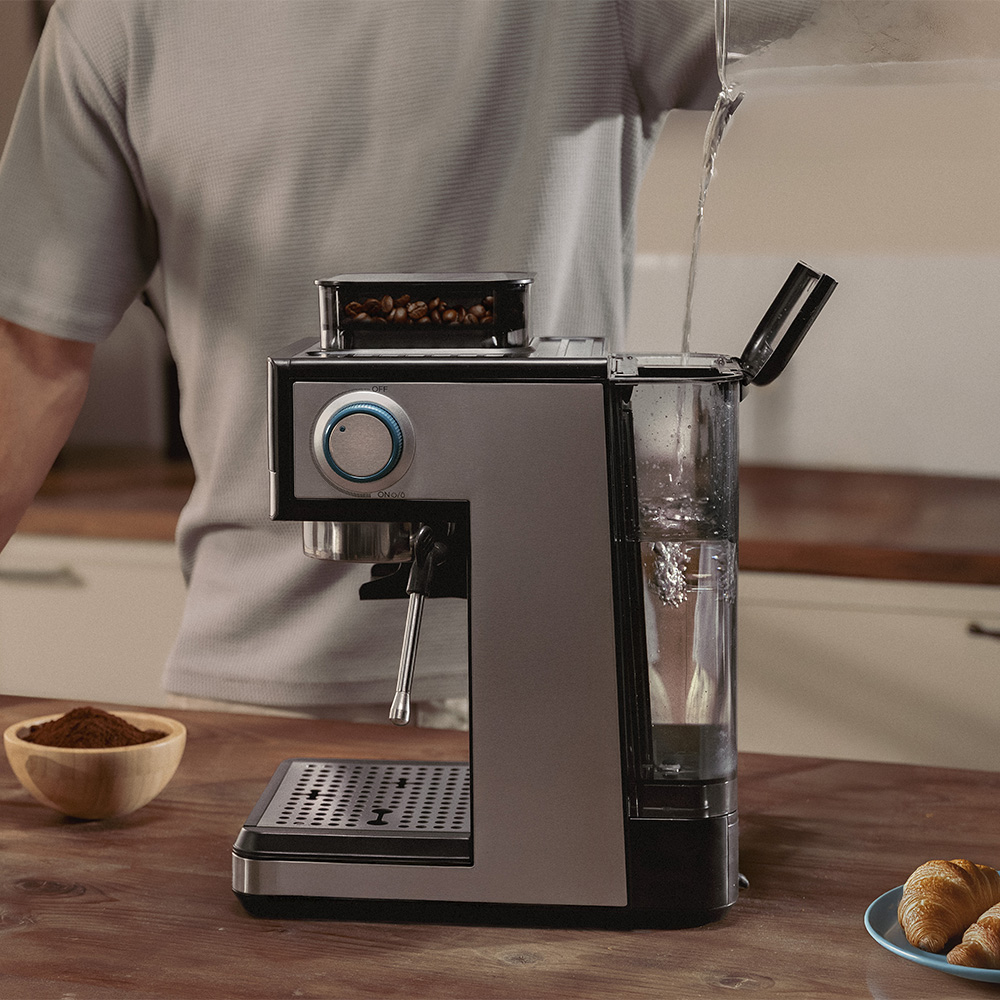 Una persona vierte agua en una cafetera. La máquina es plateada y negra, con granos de café y café molido visibles.