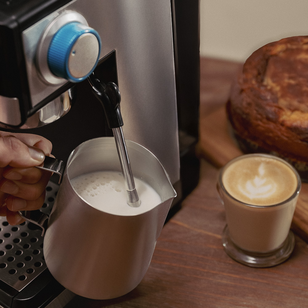 Una persona vaporiza leche con una máquina de espresso. Un café con leche y un pastel están sobre una mesa de madera. La leche espumosa está en una jarra de metal.