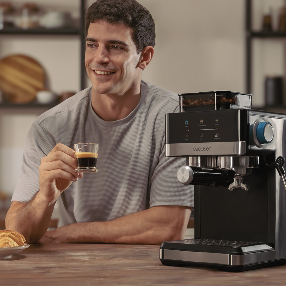 Un hombre sonríe, sosteniendo un espresso. Una cafetera negra está a su lado. Croissant en un plato sobre la mesa de madera.
