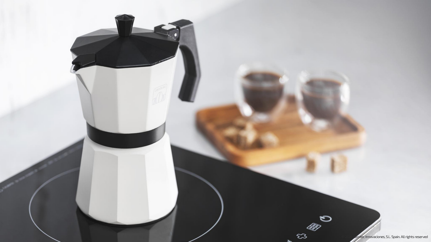 Una cafetera blanca y negra está sobre una estufa negra. Dos tazas de café y cubos de azúcar están en el fondo borroso.