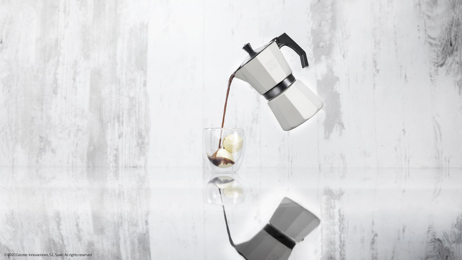 Una cafetera moka blanca vierte café sobre helado en un vaso. El fondo es una superficie blanca y texturizada. Reflejo abajo.