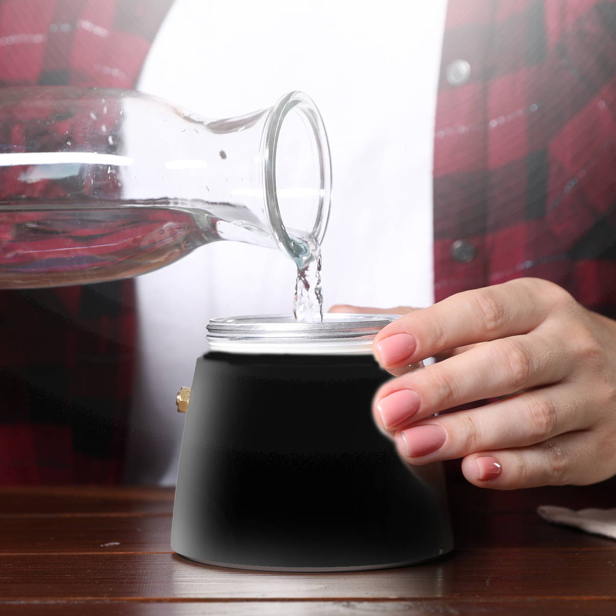 Una persona vierte agua de una jarra de vidrio en una cafetera negra. La persona lleva una camisa a cuadros roja.