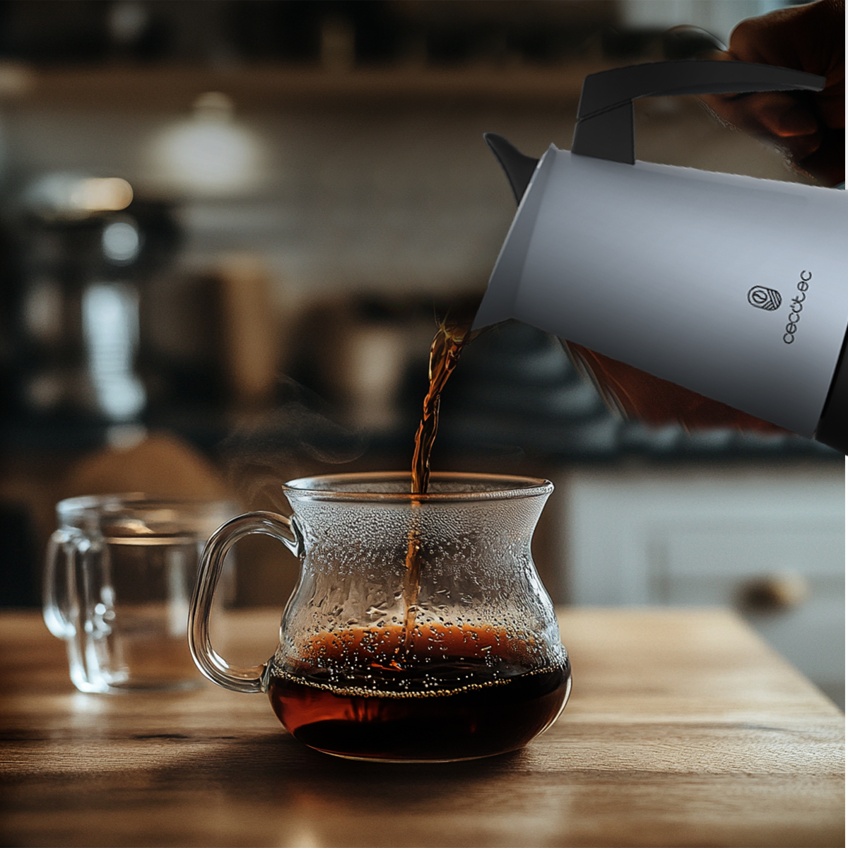Una persona vierte café de una jarra blanca en una taza de vidrio. Otra taza de vidrio vacía está en la mesa de madera.
