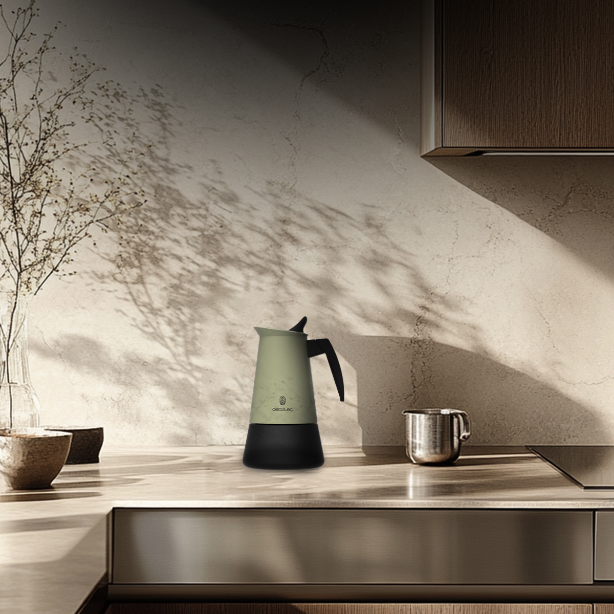 Una escena de cocina con una cafetera verde y negra. A la derecha hay una taza plateada y a la izquierda un jarrón con plantas.
