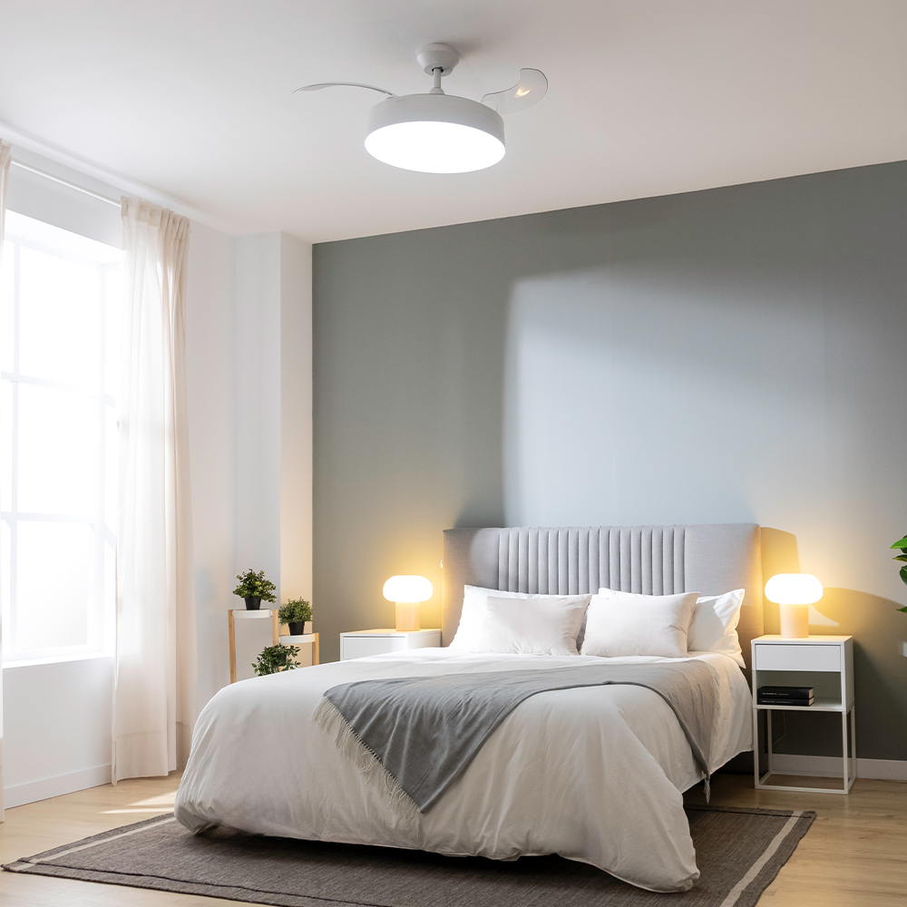 Un dormitorio con cama, mesitas de noche, lámparas y ventilador de techo. Las paredes están pintadas en un color neutro, y hay cortinas.