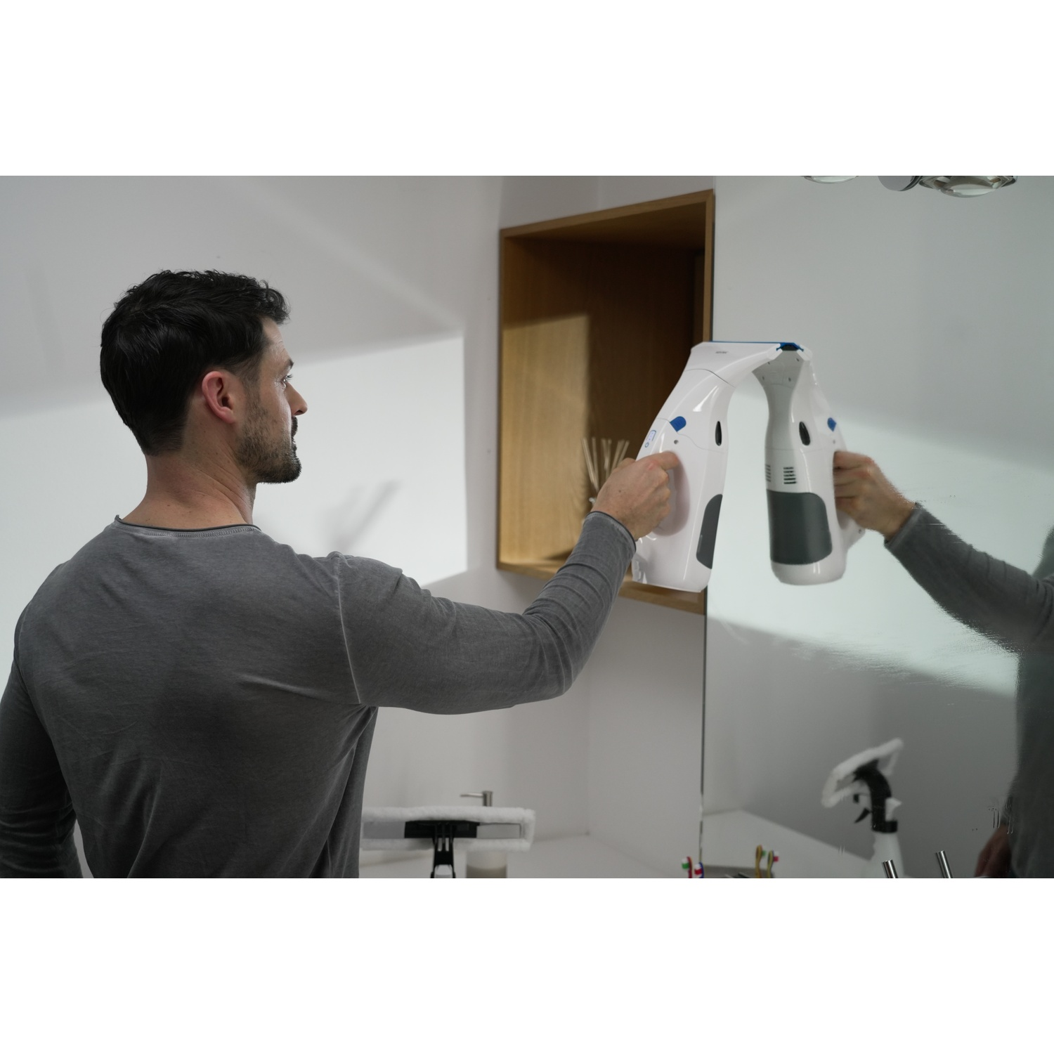 Un uomo pulisce uno specchio con un dispositivo di pulizia bianco e grigio. Il riflesso del dispositivo e dell'uomo è visibile. Un armadio sullo sfondo.