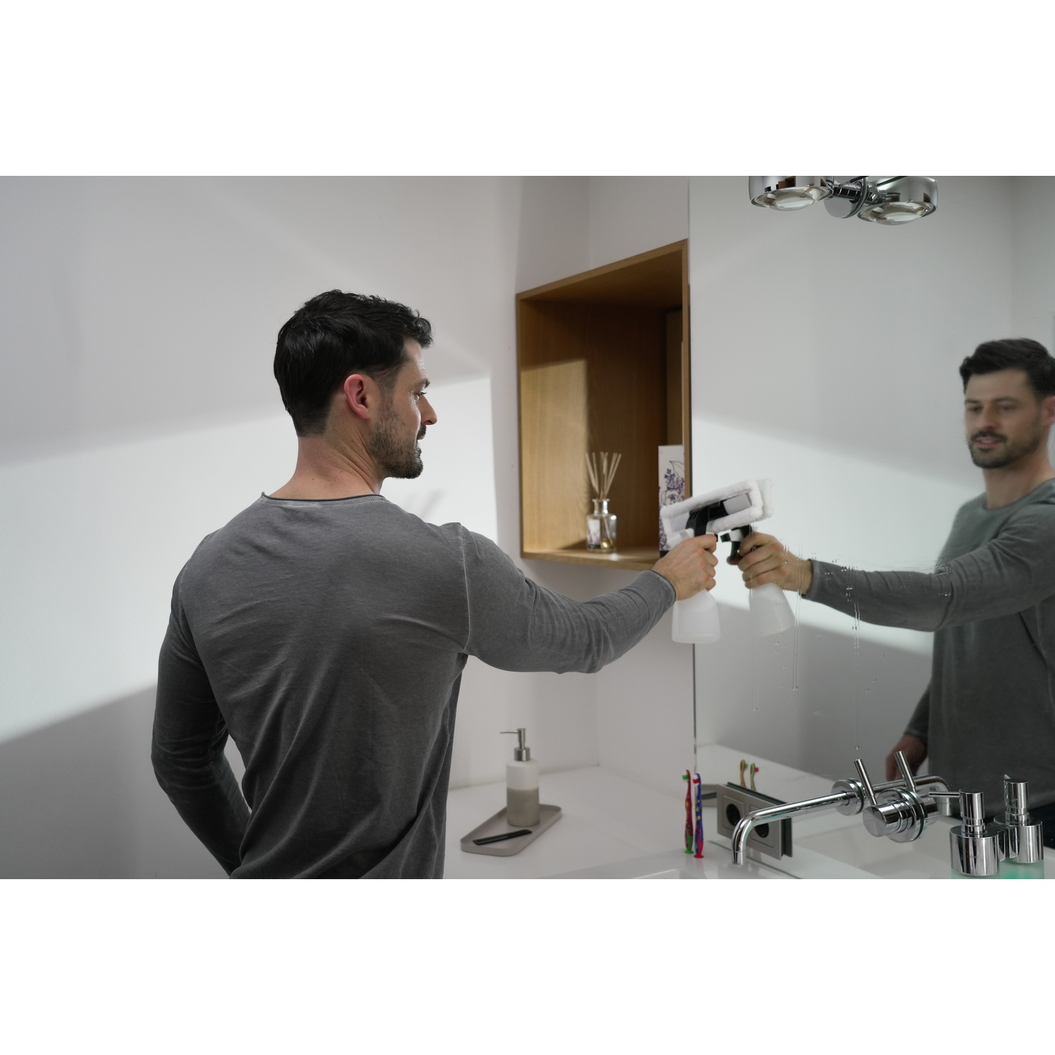 Un uomo pulisce uno specchio del bagno con un detergente spray. Lo specchio riflette l'uomo e gli accessori del bagno.
